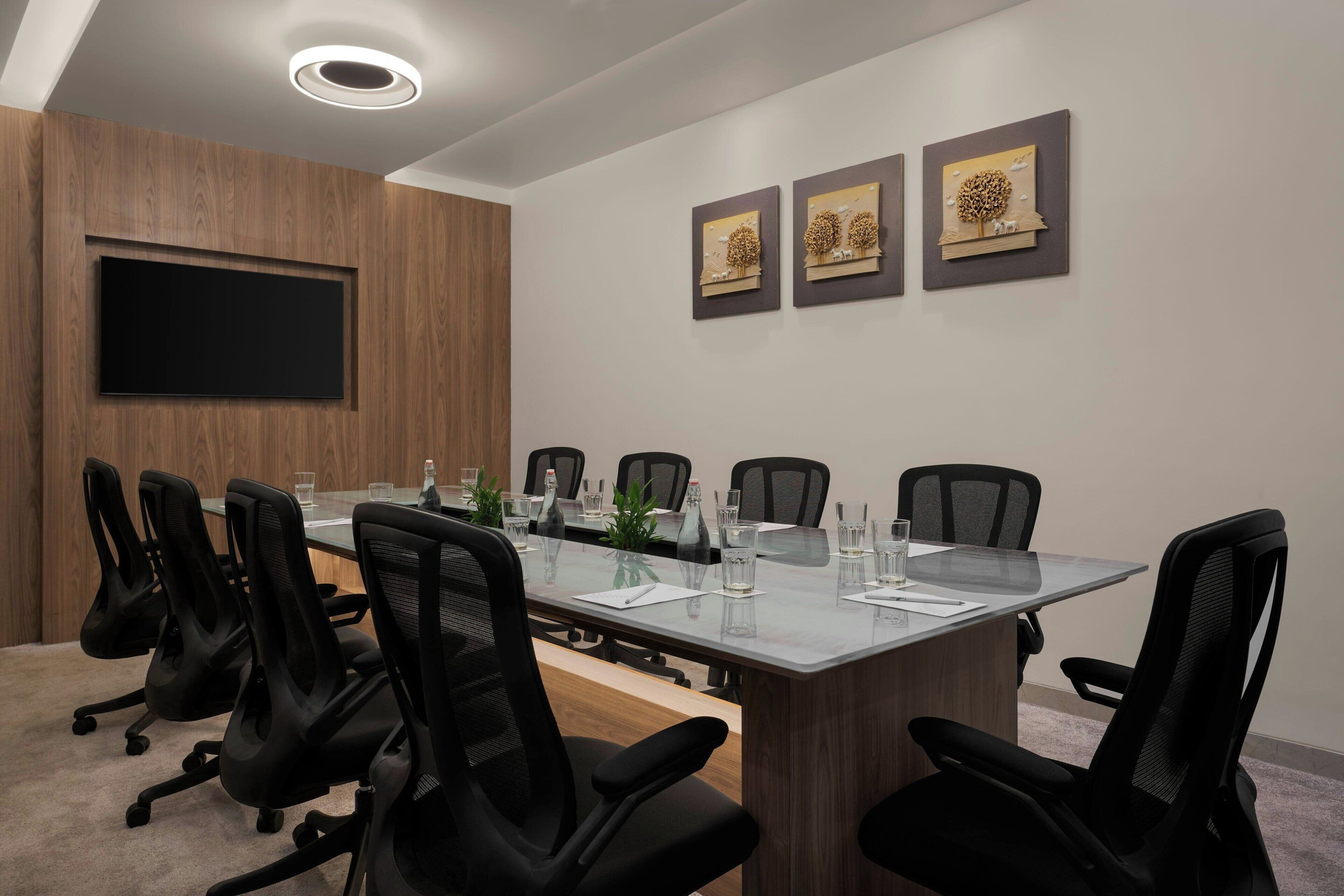 A conference room with a long table, black chairs, a flat-screen TV, and three framed artworks on the wall.