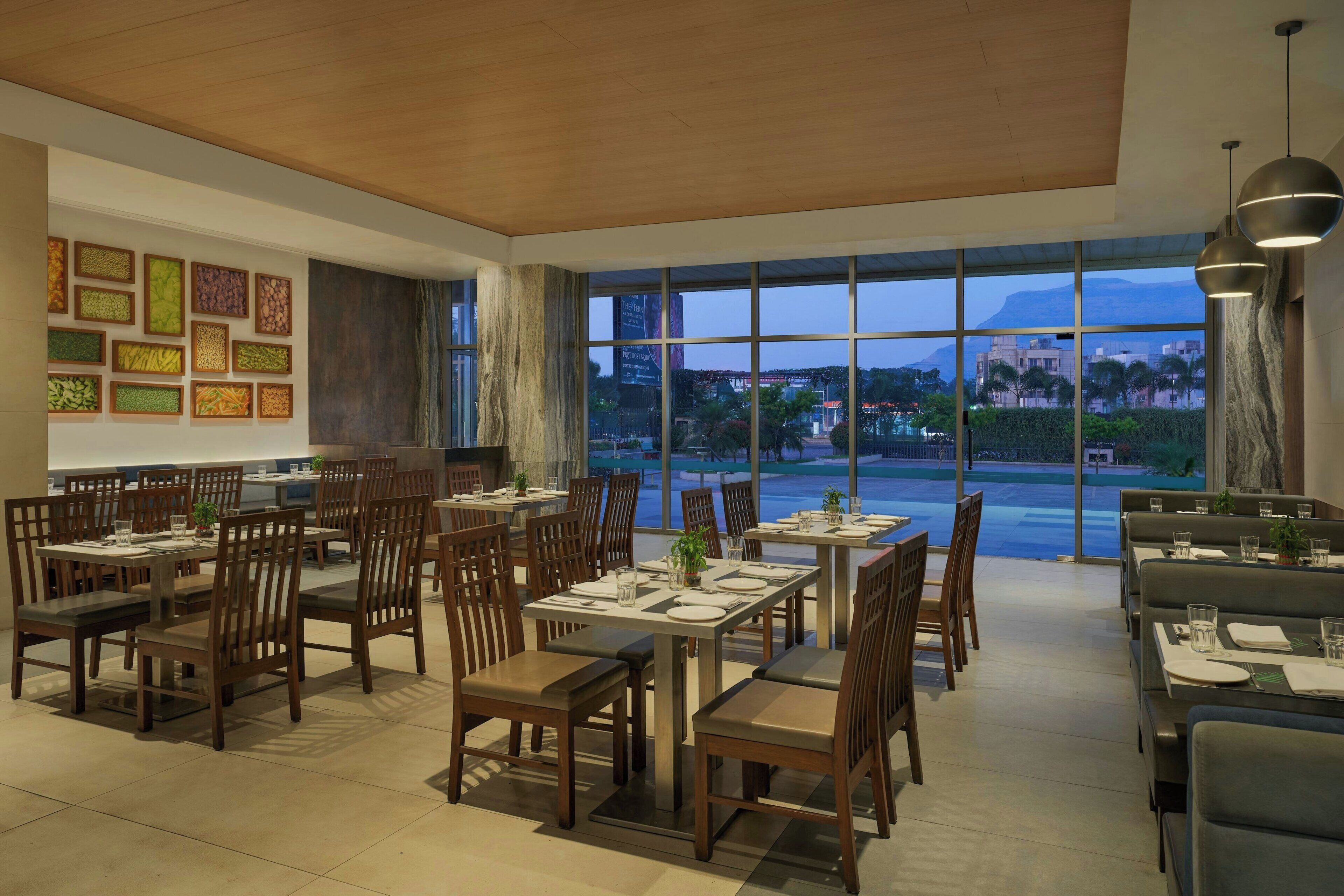 A restaurant with wooden tables and chairs, large windows with a view of a cityscape and mountains, and framed artwork on the walls.