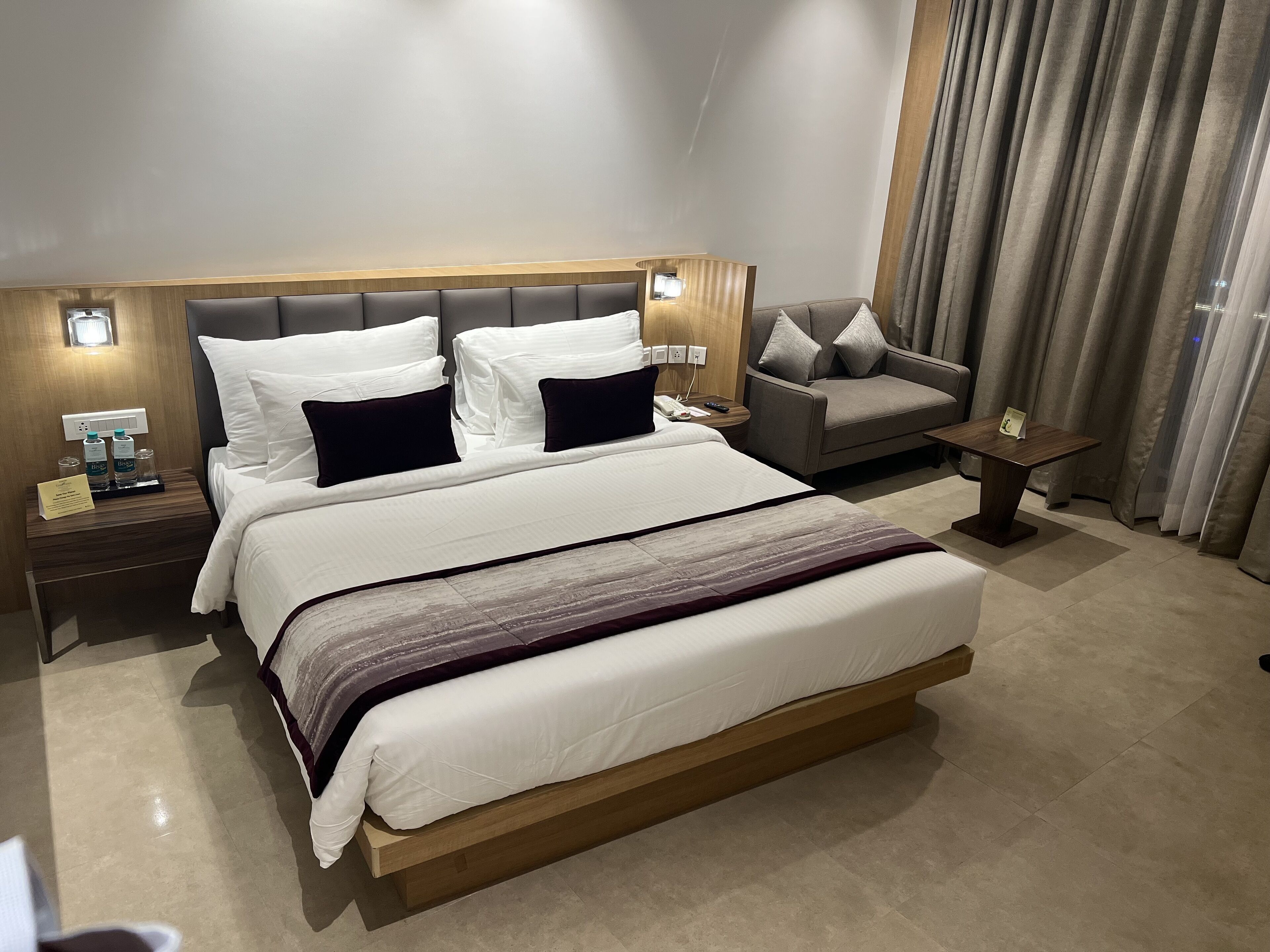 A modern hotel room with a large bed, a bedside table with a lamp, a sofa, and a small table.