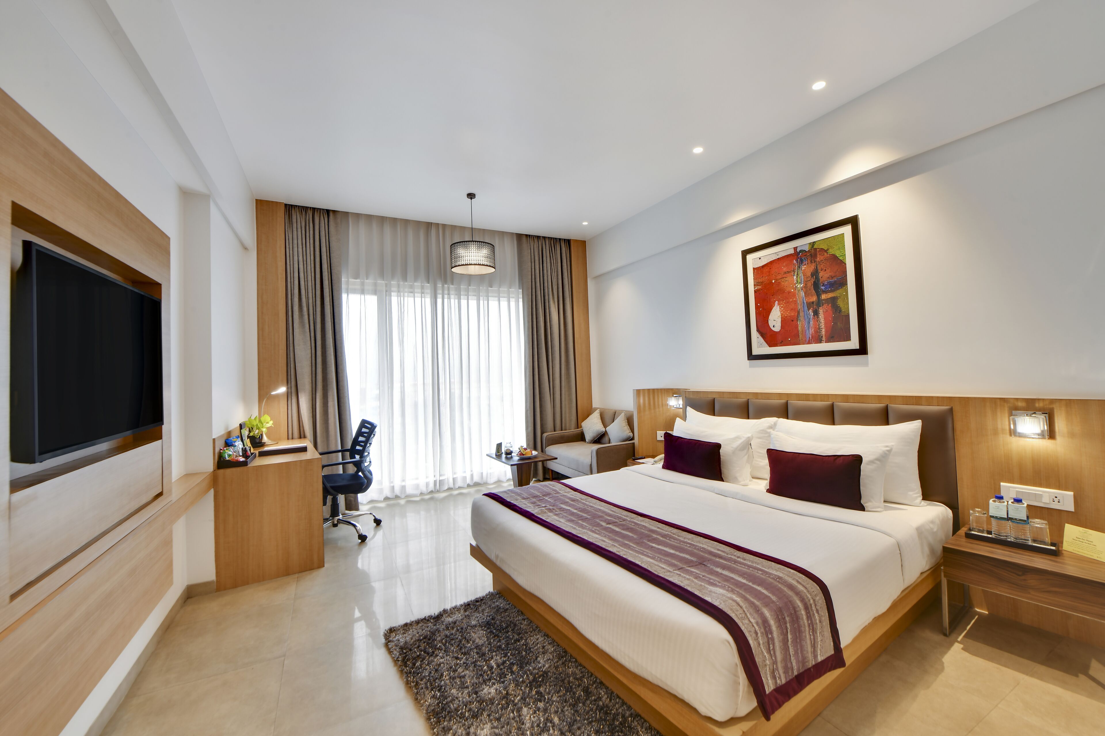 A modern hotel room with a large bed, a desk, a television, and a large window with curtains.