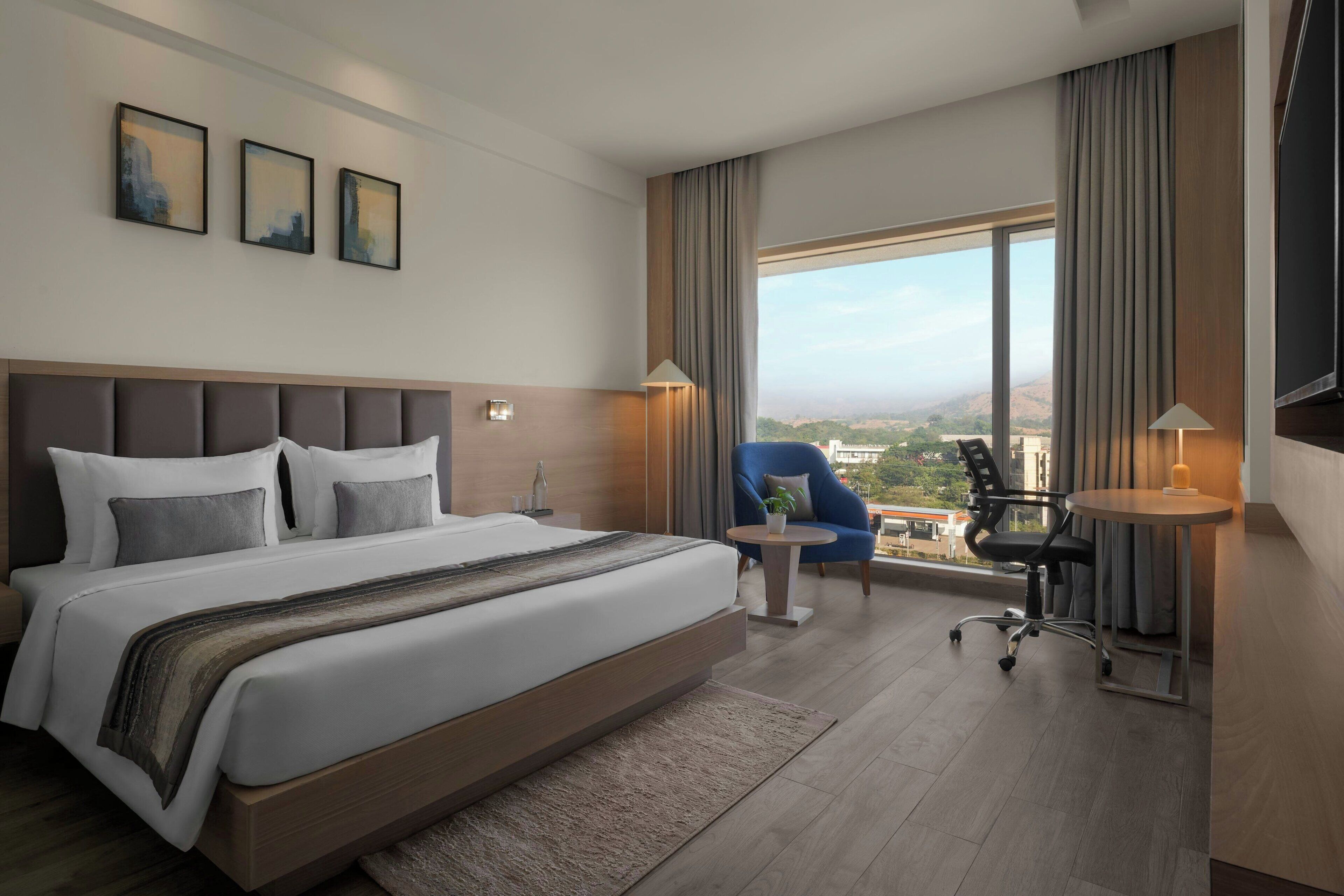 A modern hotel room with a large bed, a desk, a chair, and a view of the city.