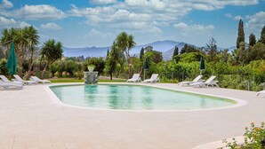 Une piscine entourée de chaises longues blanches et agrémentée d’une fontaine en pierre, le tout niché au cœur d’un écrin de verdure, avec des montagnes en toile de fond.