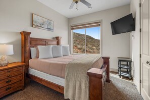A bedroom with a large bed, a nightstand, a dresser, a TV mounted on the wall, and a window with a view of a hilly landscape.