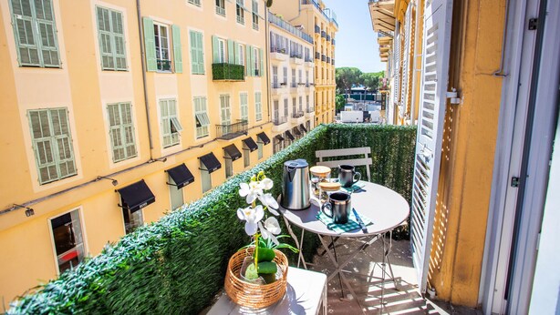 A balcony with a table set for two, a wicker basket with flowers, and a view of adjacent buildings. - PARADISIO 2 AP4237 By Riviera Holiday Homes (Niza)