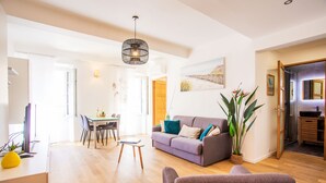 A modern living room with a grey sofa, a dining area with a table and chairs, a large potted plant, and a kitchen area visible through an open door. - PARADISIO 2 AP4237 By Riviera Holiday Homes (Niza)