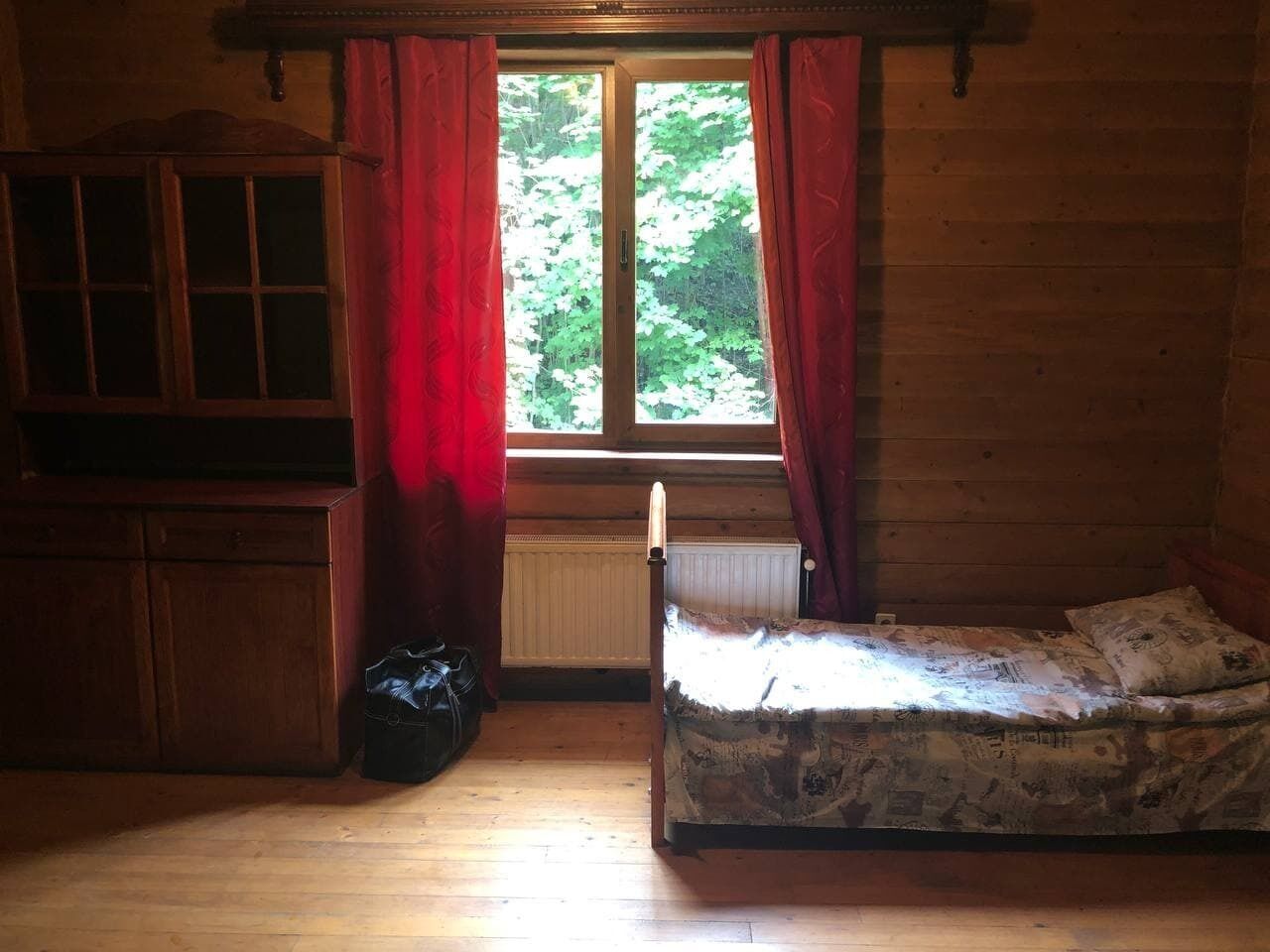Een kleine, zwak verlichte kamer met een enkel bed, een houten kast en een raam met rode gordijnen.