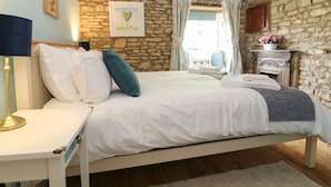 A bedroom with a stone wall, a bed with white bedding, a bedside table with a lamp, and a small table with a flower vase. - Tuesday Cottage (Cheltenham)