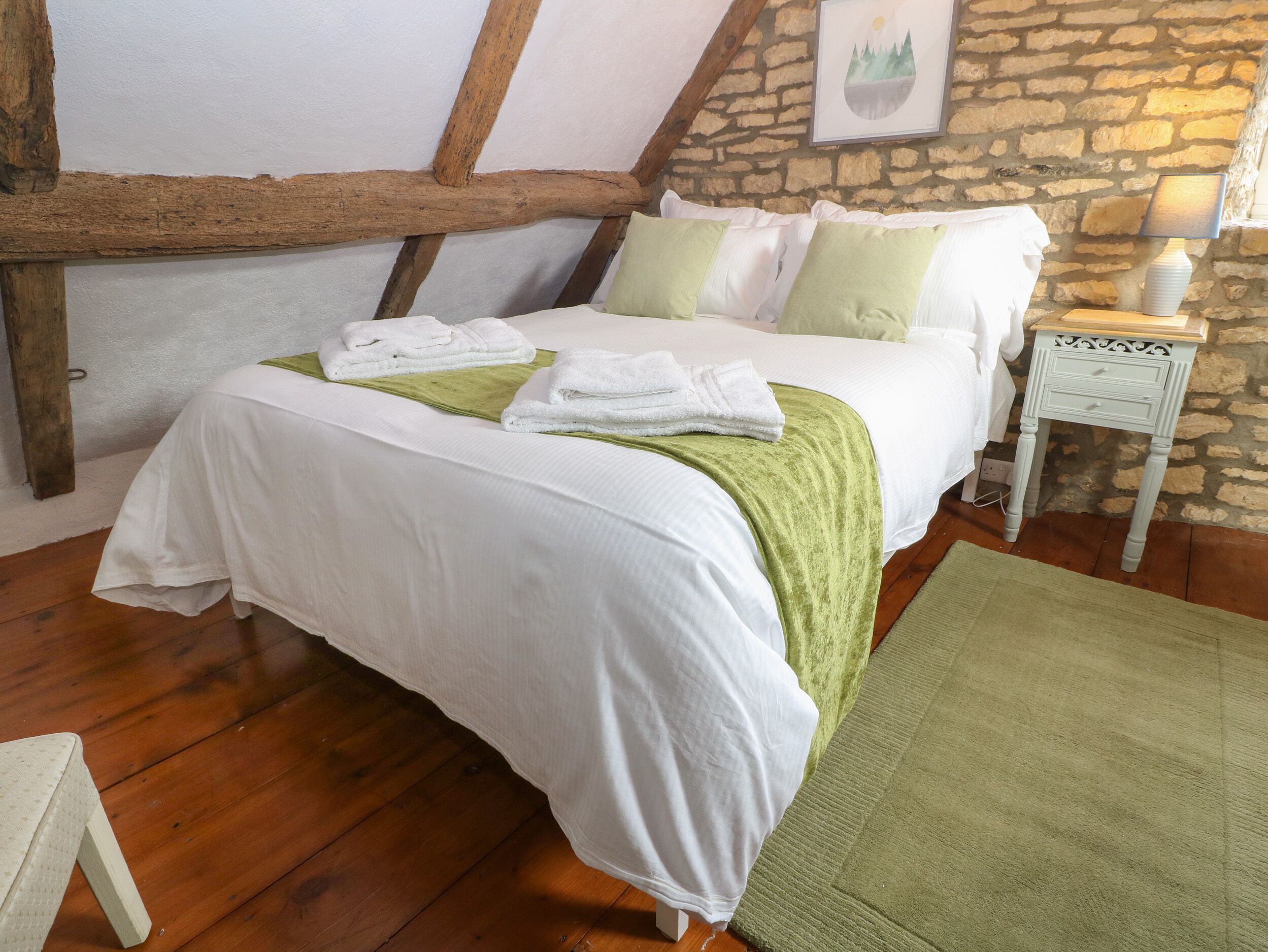 A neatly made bed with white linens and green accents, a stone wall, and a wooden floor.