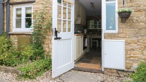 A stone cottage with a sign reading "Tuesday Cottage." It features a kitchen with a window, a hanging plant, and a door leading inside. - Tuesday Cottage (Cheltenham)