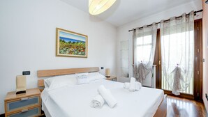 Ein Hotelzimmer mit einem ordentlich bezogenen Bett, einem Nachttisch mit Lampe, einem Fenster mit Vorhängen und einem Bild an der Wand.