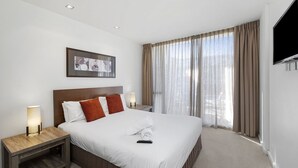 A hotel room with a large bed, two bedside tables, a TV mounted on the wall, and a window with curtains. - Resort Apartment 224 (TORQUAY)