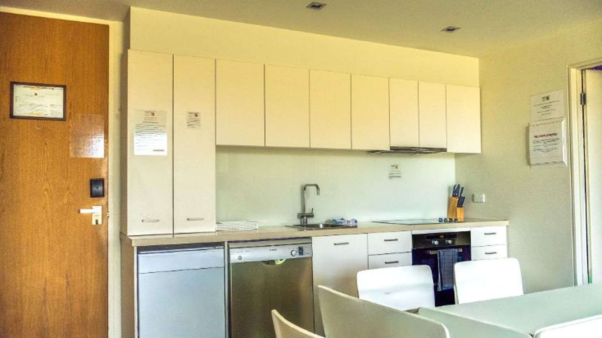 A compact kitchen with white cabinets, a dishwasher, and a sink. There are two white chairs at a table.