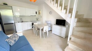 A modern kitchen with white cabinets, a dining area with a table and chairs, a sofa with a patterned pillow, a TV on a white dresser, and a staircase with white railings. - Affittimoderni Viddalba Bouganville - VIBO09 (Viddalba)