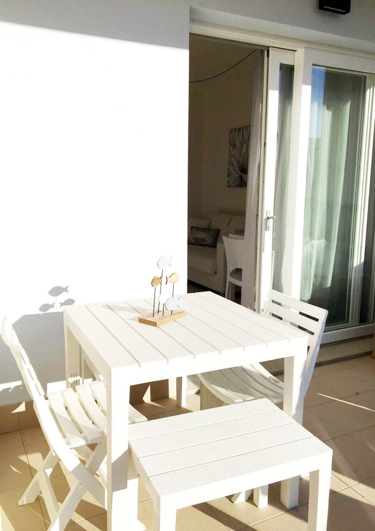 Een witte eethoek voor buiten met een tafel en bankjes, een klein decoratief object op de tafel, en een zicht op het interieur door een glazen deur.