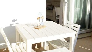 A white outdoor dining set with a table and benches, a small decorative piece on the table, and a view of the interior through a glass door. - Affittimoderni Viddalba Bouganville - VIBO09 (Viddalba)