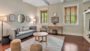 Ein Wohnzimmer mit grauem Sofa, rundem Couchtisch und zwei Hocker. Es gibt zwei Fenster mit Jalousien, einen Schreibtisch und eine Lampe.