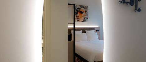 A modern hotel room with a large oval mirror, a bed with white linens, a headboard, and a painting on the wall.