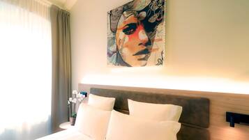 Une chambre d’hôtel avec un lit, des oreillers blancs, une tête de lit, une table de chevet avec une lampe, une fenêtre avec des rideaux et un tableau au mur.
