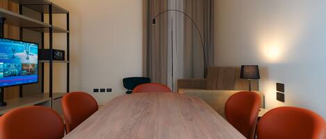 A conference room with a wooden table, orange chairs, a television on a shelf, a lamp, and a window with curtains.