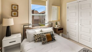 Quarto com cama, mesa de cabeceira, abajur, cômoda e uma janela grande com vista para casas.