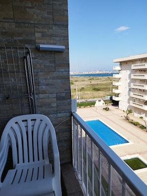 A balcony with a white plastic chair, a stone wall, and a view of a swimming pool and distant buildings.