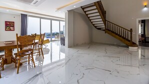 A spacious room with a marble floor, a dining area with wooden chairs, a staircase with wooden railings, and large windows offering a view.