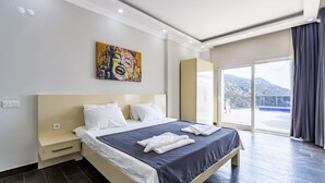 A hotel room with a large bed, a nightstand, a painting on the wall, and a view of the mountains through the window.