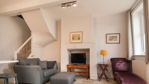Un salon avec un canapé gris, un fauteuil violet, un téléviseur et un escalier.