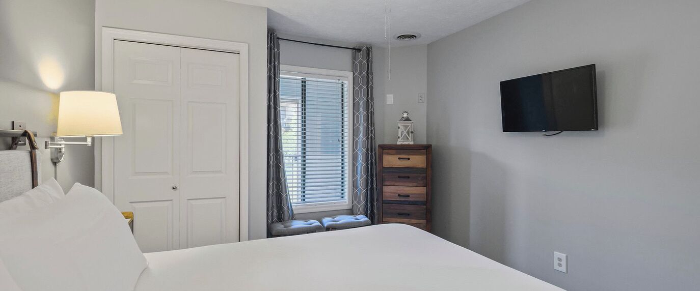Une chambre d’hôtel avec un lit, un téléviseur fixé au mur, une fenêtre avec des stores, une lampe de chevet et une commode en bois.