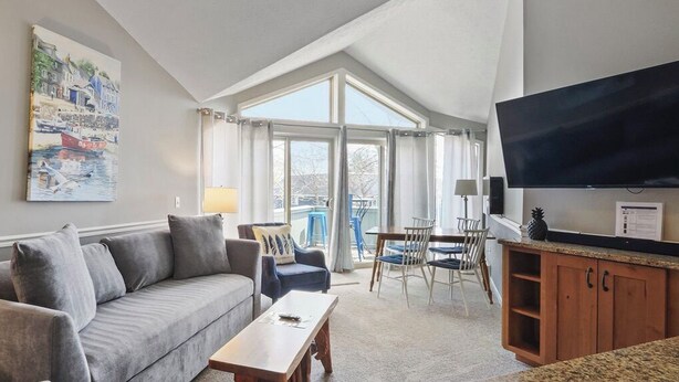 A living room with a sofa, coffee table, and TV. There is a balcony with a table and chairs. - 211 Pointes North | Dog Friendly | Downtown Charlevoix | Indoor/Outdoor Pool (Charlevoix)