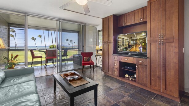 A modern kitchen with a large TV, wooden cabinets, and a sliding glass door leading to a balcony with a view of palm trees. - Leinaala Oceanfront #203 Panoramic Ocean View, Renovated, A/C, Great Rates! (Kihei)