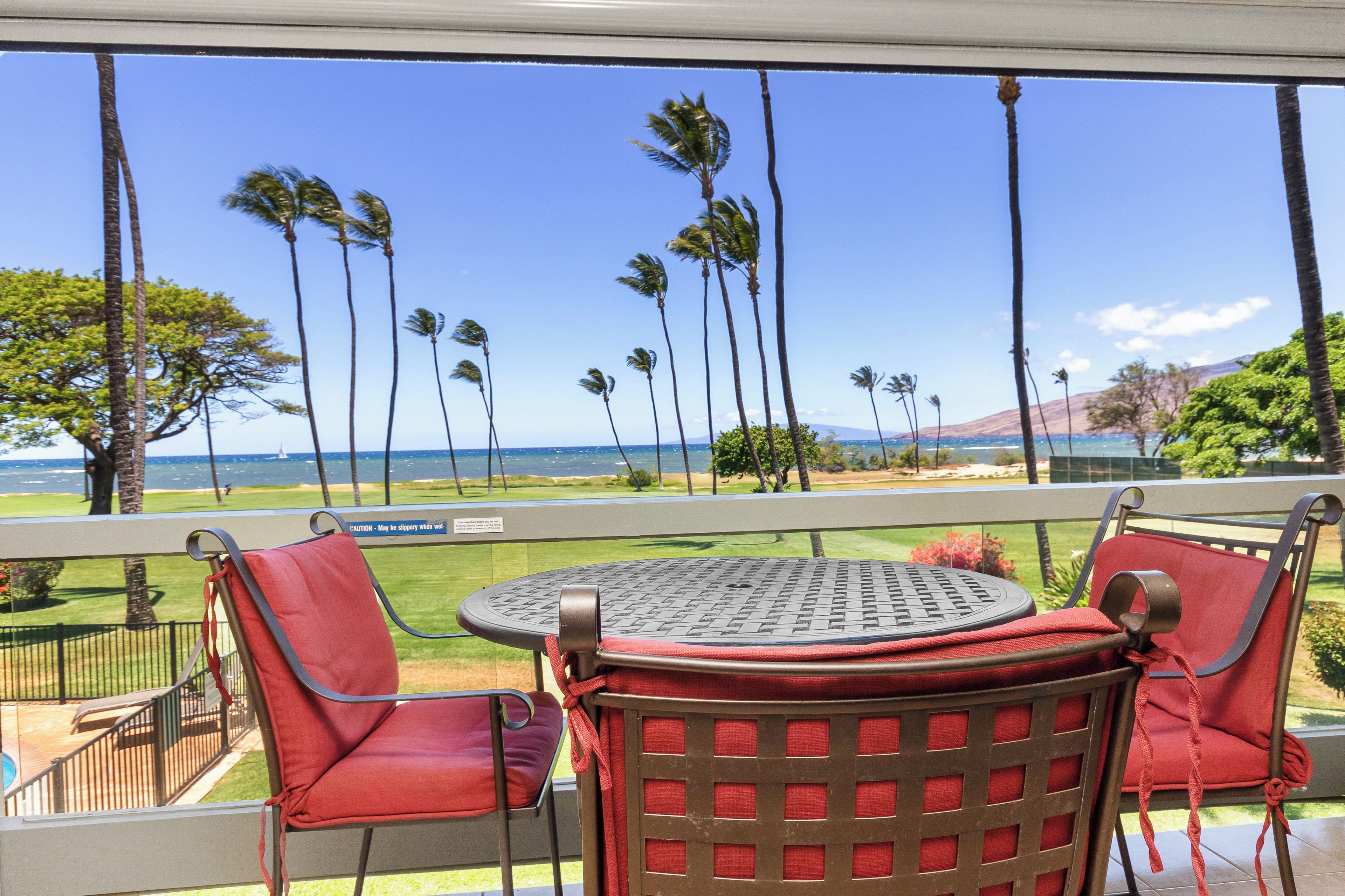 A balcony with red chairs and a round table, overlooking a view of palm trees and the ocean.