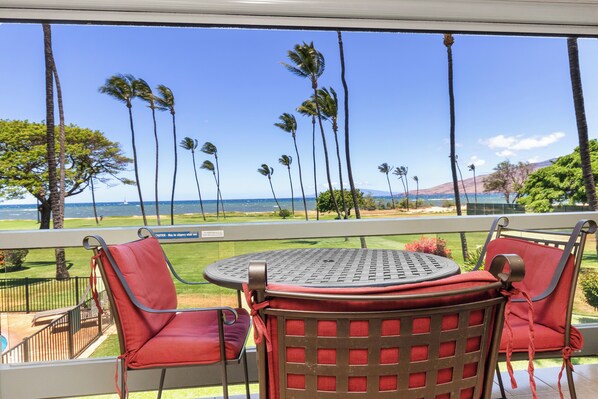 A balcony with red chairs and a round table, overlooking a view of palm trees and the ocean.