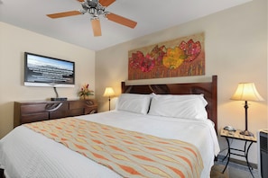 A hotel room with a bed, a ceiling fan, a television, a painting, and two bedside lamps.