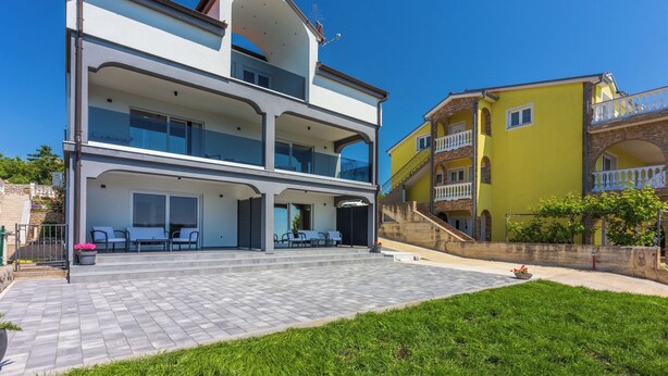 En toetagers bygning med en overdækket terrasse, en trappe op til en balkon og et lille haveområde.