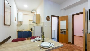 Ένας χώρος τραπε ζαρίας με ένα ξύλινο τραπέζι στρωμένο για δύο άτομα, μια κουζίνα στο βάθος και μια πόρτα που οδηγεί σε ένα άλλο δωμάτιο.