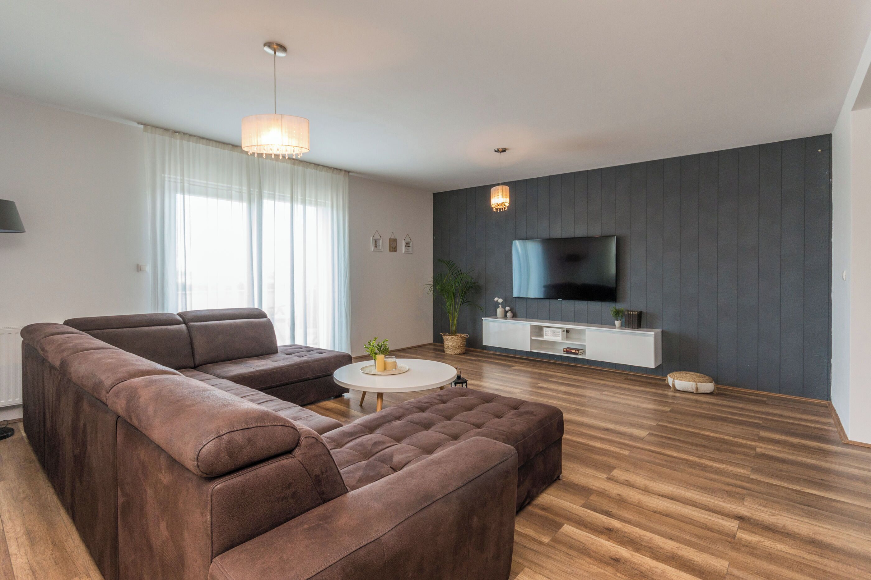 A modern living room with a sectional sofa, a round coffee table, and a flat-screen TV mounted on a dark wall.