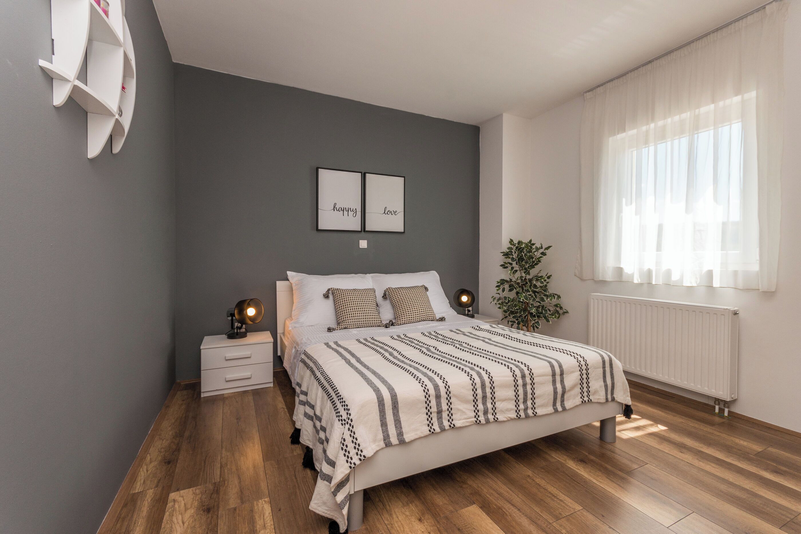 Quarto com cama, mesas de cabeceira, prateleira na parede, planta em vaso e janela com cortinas.