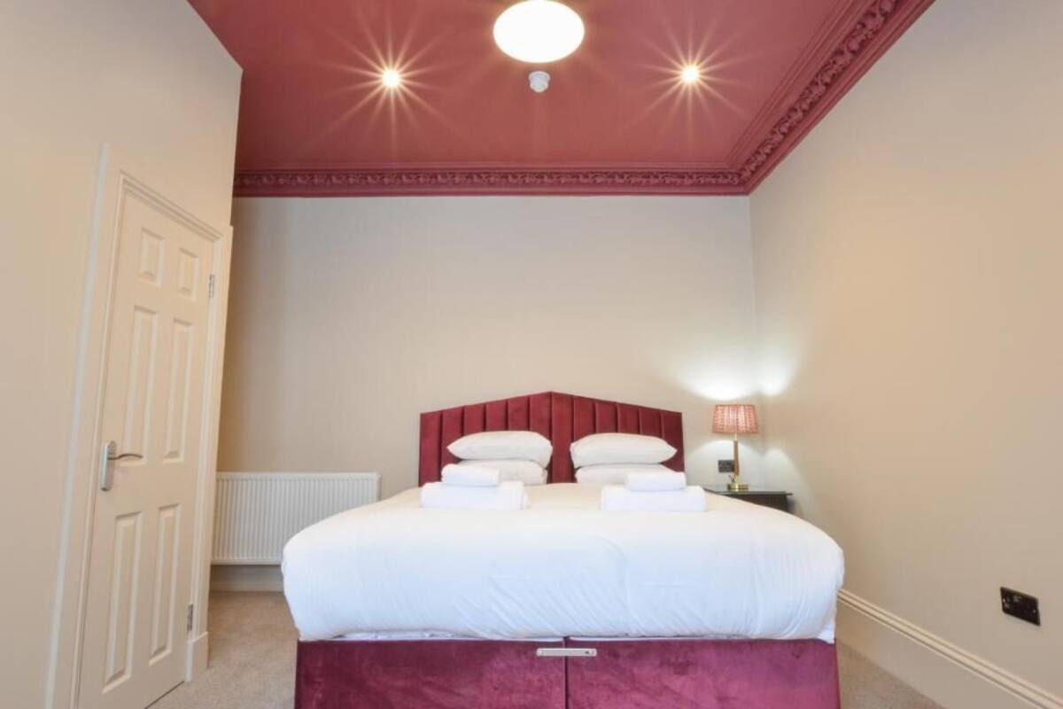 A bedroom with a large bed, a red headboard, a nightstand with a lamp, and a white door.