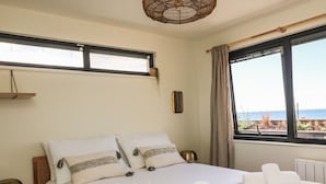 A neatly made bed with white linens and decorative pillows, a bedside lamp, and a view of the sea through a large window. - Sandpiper Chalet (Bridport)