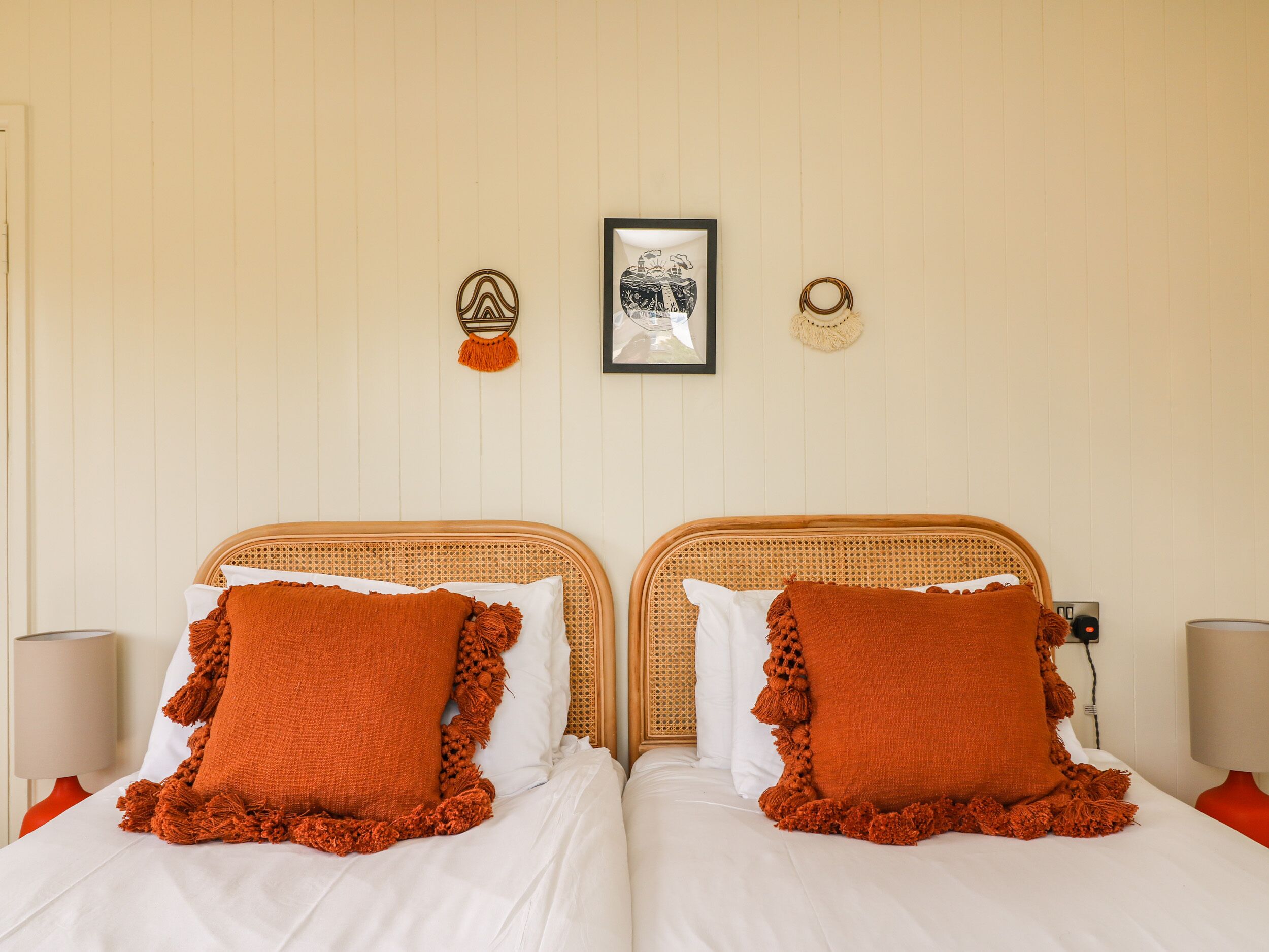 A double bed with white bedding and orange pillows, a framed picture above the bed, and a wall-mounted decorative item.
