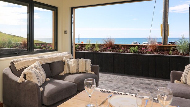 A modern living room with a large window overlooking a scenic view of the sea and a parking area. - Sandpiper Chalet (Bridport)
