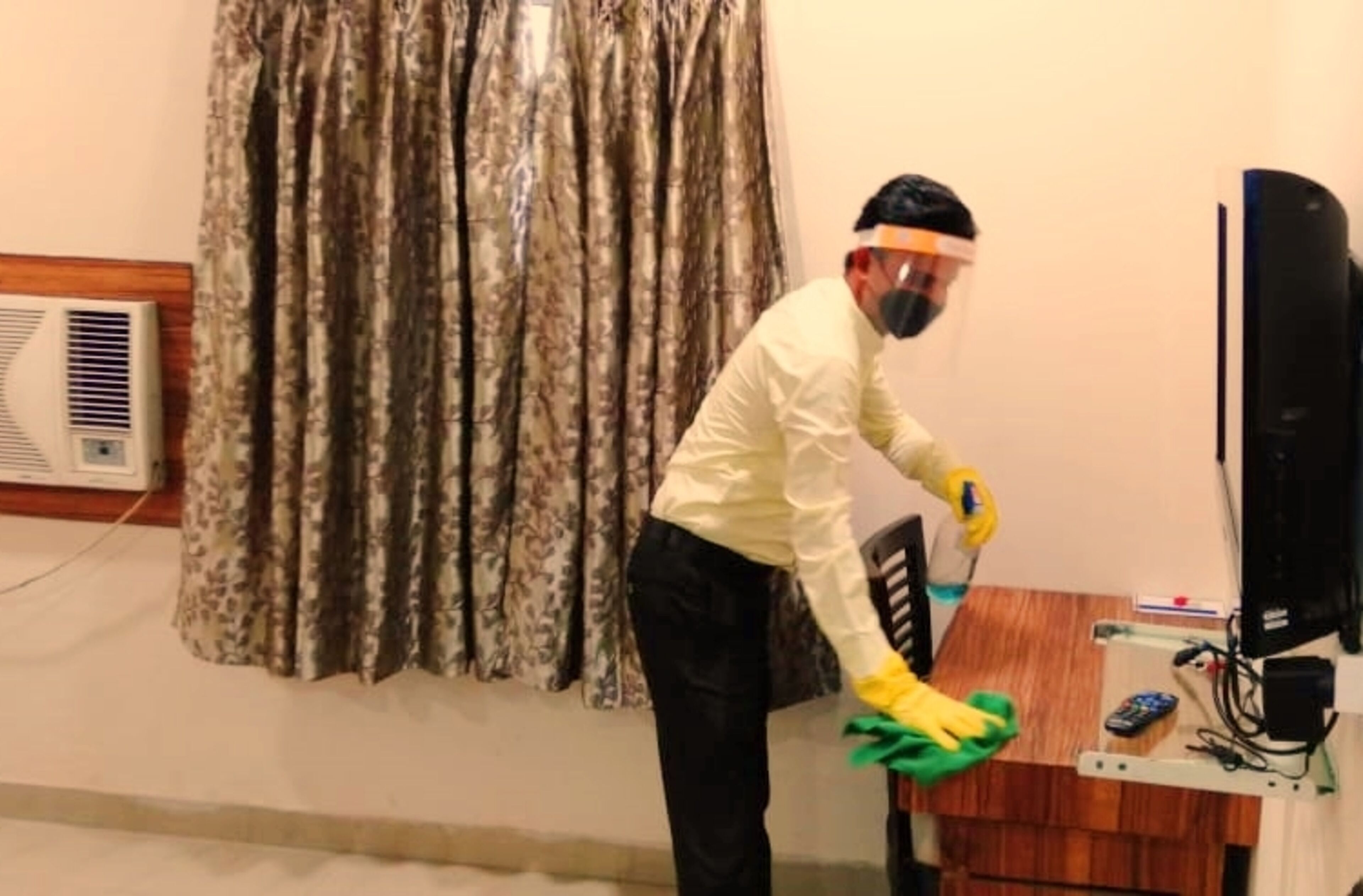 A person cleaning a wooden table in a room with a curtain, a wall-mounted air conditioner, and a TV.
