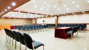 Un salón de conferencias con filas de sillas, una mesa con mantel rojo y un ventilador de techo.