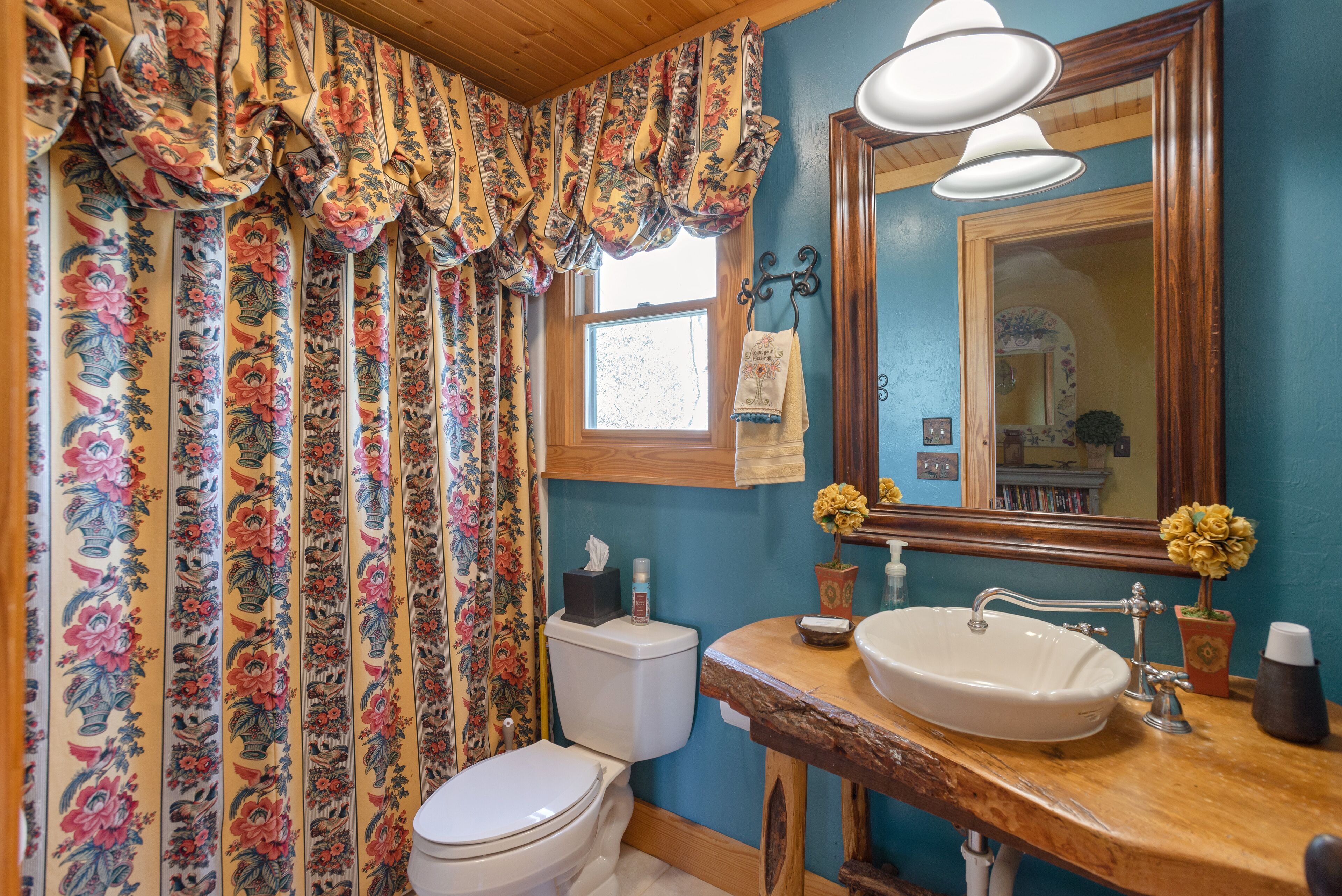 A bathroom with a floral curtain, a white toilet, a wooden vanity with a sink, and a mirror.