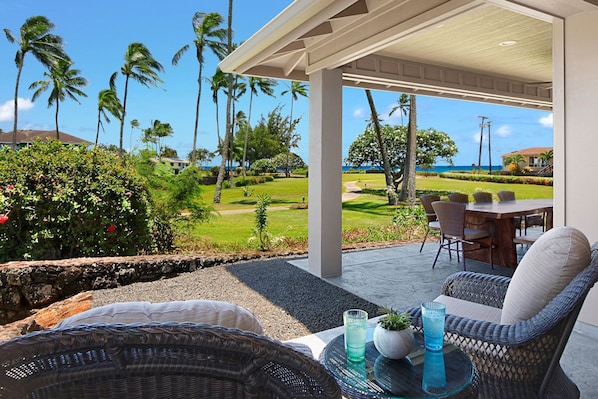 A covered outdoor seating area with wicker furniture, a glass table, and a view of a golf course and ocean.