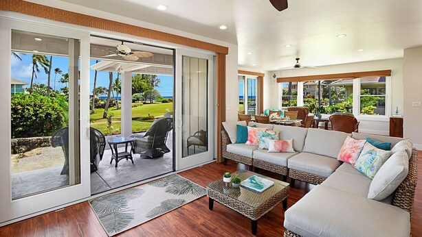 A spacious living room with a large sliding glass door leading to a patio, a ceiling fan, and a view of a lush garden.