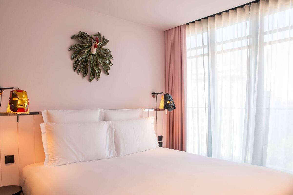 A hotel room with a bed, a wall-mounted plant, and a window with curtains.