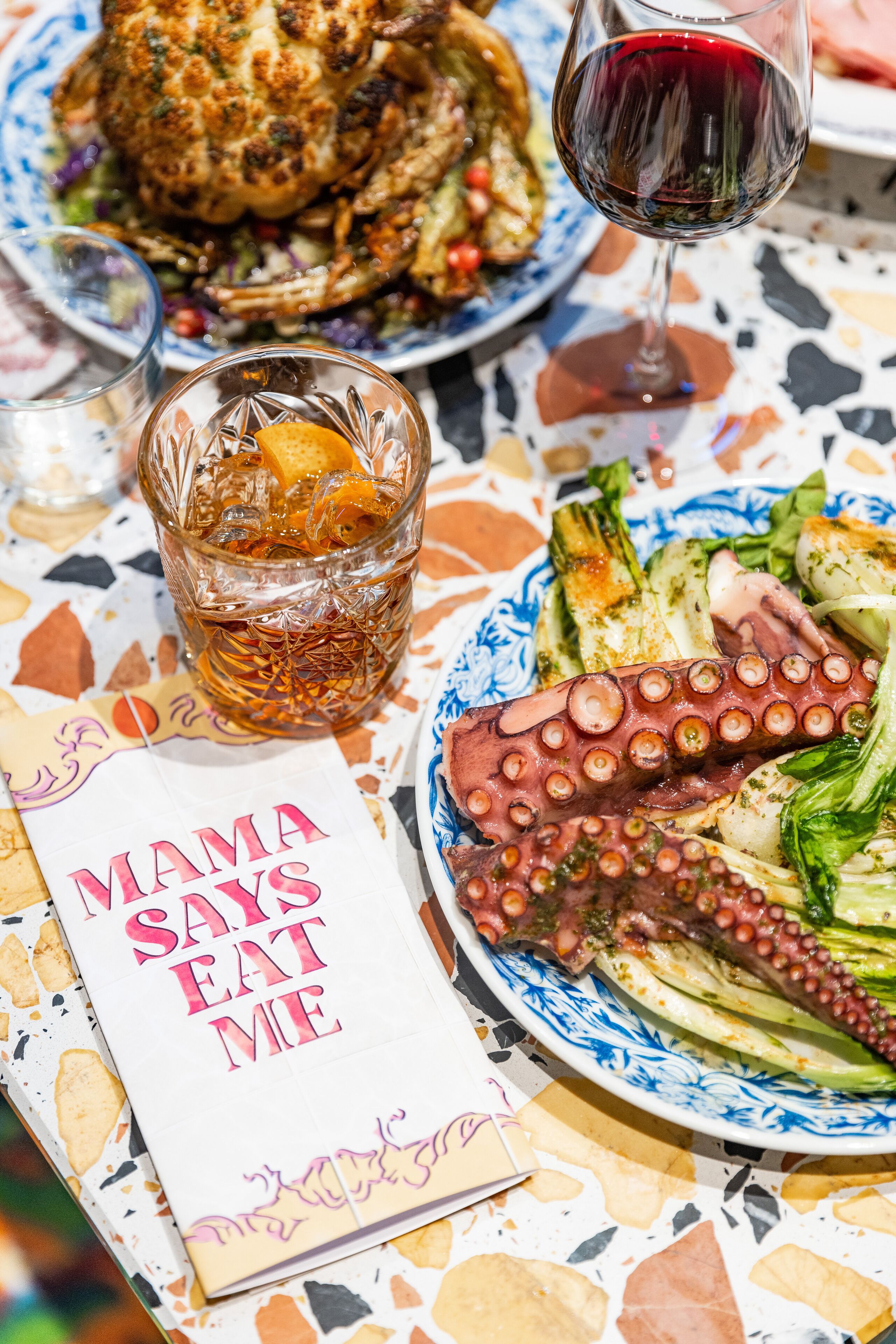 Een eettafel met een bord octopus, een glas rode wijn en een menukaart met de tekst 'MAMA SAYS EAT ME'.
