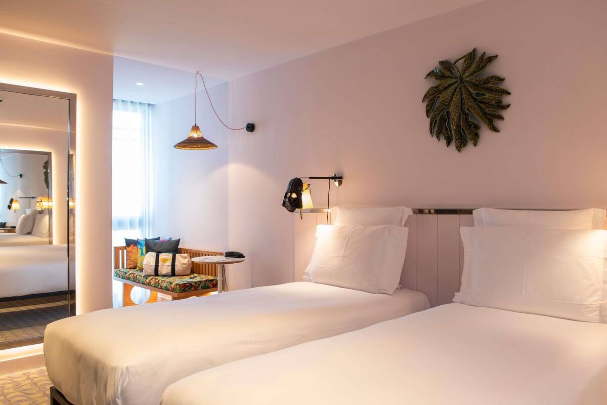 A hotel room with two beds, a small table with a chair, a wall-mounted lamp, and a decorative leaf on the wall.